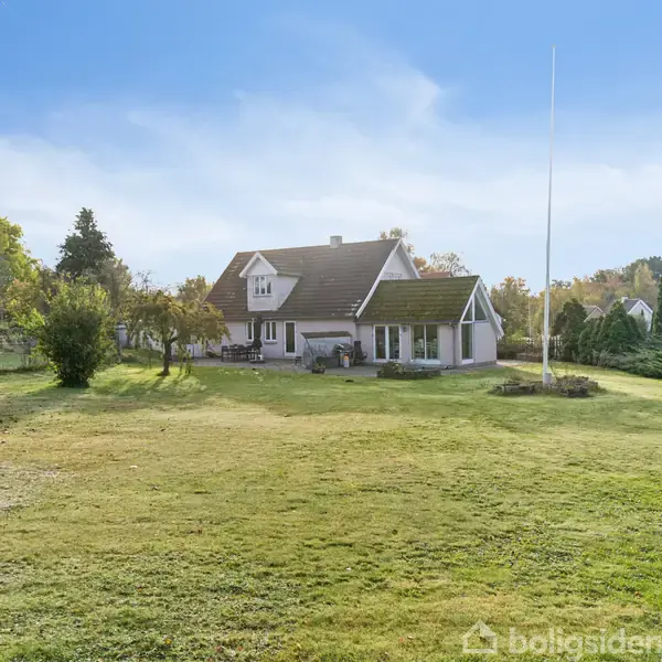Et hvidt hus med sort tag står på en velplejet græsplæne, omgivet af træer og buske. Himlen er blå, og der ses en flagstang i nærheden.