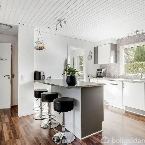 Køkken med hvidt interiør har en central ø med tre barstole, planter på bordet, og vindue med udsigt til have. Dør viser teksten "Velkommen".