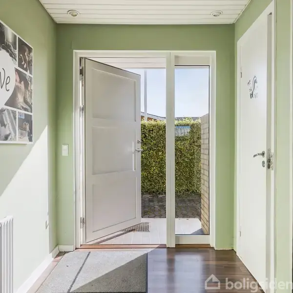 Dør står åben i en entré med grønne vægge. Et fotocollage med teksten "Love" hænger på venstre væg. Udenfor ses grønne hække og blå himmel.