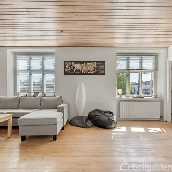 En grå sofa står foran to store vinduer, omgivet af en trævæg og trægulv. En stor lampe og to sækkestole står ved siden af. Værelser lyses op af naturligt lys.