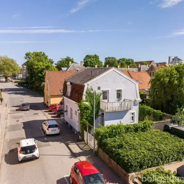 Et hvidt hus med balkon ligger på en rolig villavej omgivet af grønne træer og hække. Flere biler kører og parkerer langs vejen, under en klar blå himmel.