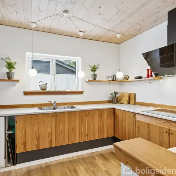 Køkken med træskabe og hvide bordplader, udstyret med køkkenvask og kogeplade. To åbne hylder med planter og krukker, belyst af hængelamper. Træloft og lyse trægulve.