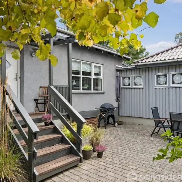 Et terrasseområde med hegn og trapper fører op til et gråt hus. Blomsterkrukker står spredt rundt omkring, og der er havemøbler samt en grill i baggrunden. Træerne omkranser terrassen.