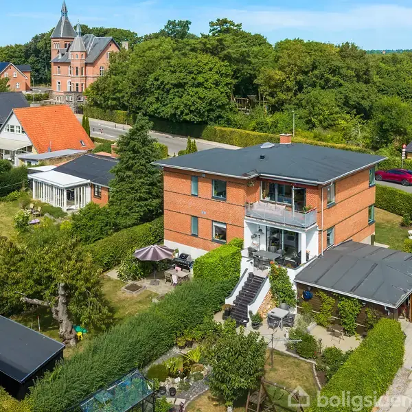 En rød murstensbygning med altan, omgivet af en velplejet have med stier og buske. Bygningen har en trappe, der fører til en terrasse. Naboer og træer omkranser området.