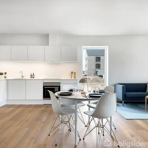 Et lyst og moderne køkken-alrum med hvide skabe, en indbygget ovn og komfur. Et rundt spisebord med stole står i midten. I baggrunden ses en blå sofa med bord.
