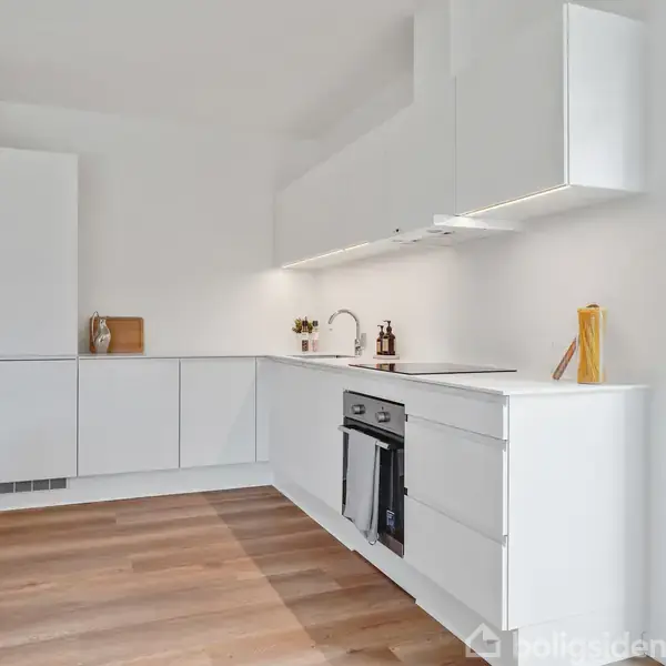 Hvidt moderne køkken med indbygget ovn og komfur, minimalistiske skabe og en vask. Trægulv dækker gulvet. Rummet er lyst med naturligt lys fra et stort vindue på højre side.