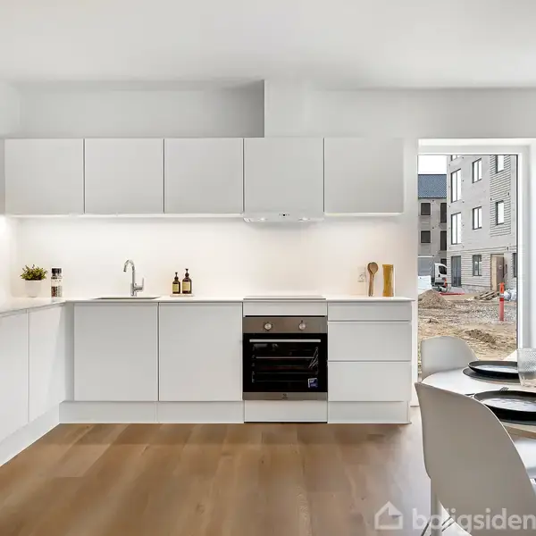 Et hvidt køkken med indbyggede skabe og en ovn. Et spisebord med sorte tallerkener ses til højre. Gennem et vindue ses en boligbygning udenfor.