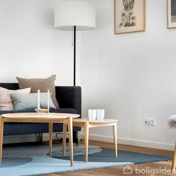 Sofa med puder står mod en hvid væg ved siden af et rundt sofabord med lys på en blå tæppe. En gulvlampe lyser rummet, der har træmøbler og kunst på væggen.