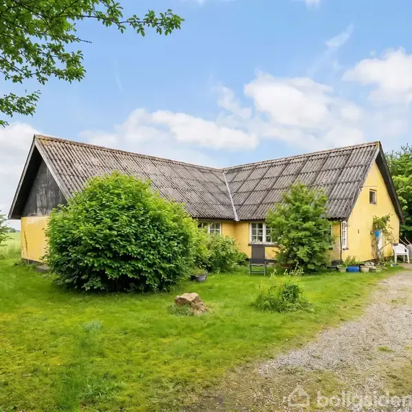 Gul landhus med tegltag står omgivet af frodige buske og en grussti i en grøn have. Himlen er blå med spredte skyer.