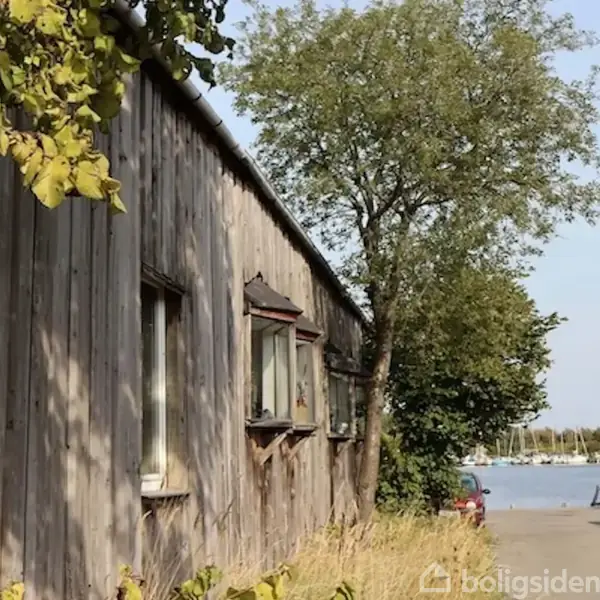 Træbygning med vinduer står langs en vej med parkerede biler og træer. I baggrunden er en marina med flere mastede både, hvilket antyder nærhed til vand.