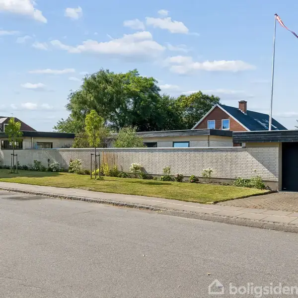 En etplans villa med en hvid murstensmur og garageport. Den står på en velholdt græsplæne med enkelte buske, omgivet af andre huse og træer langs en rolig vej.