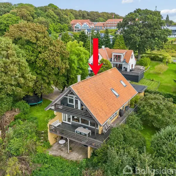 Huset ligger i et grønt område omgivet af træer, med en rød pil pegende mod det orange tag. Der er flere huse i baggrunden i et landområde.
