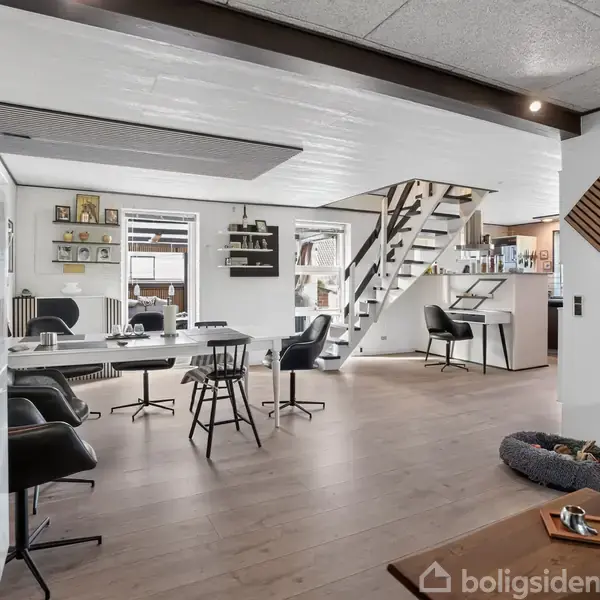 En moderne stue har et minimalistisk design med sorte og hvide møbler, inkludert stole og borde. Der er en trappe op til næste etage. Væggene er dekoreret med hylder og kunst.