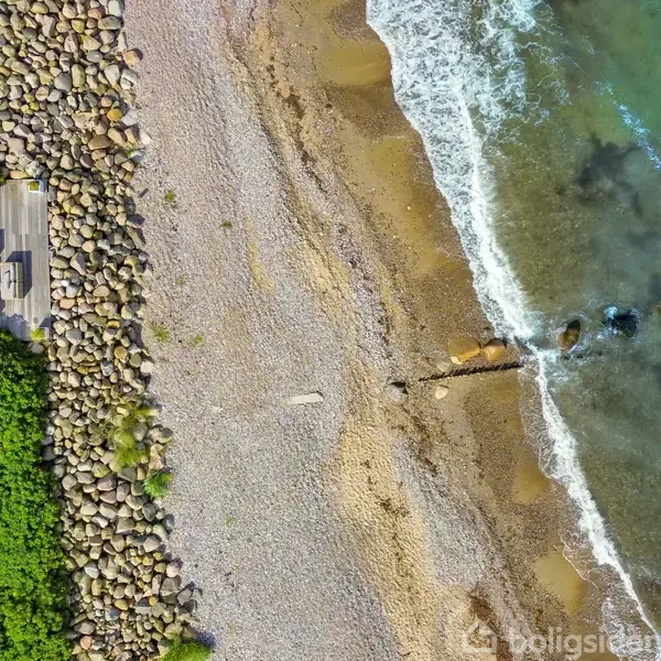 Strand ved havet med bølger. Et hus og terrasse med møbler er omgivet af grønne buske. Store sten adskiller terrassen fra stranden. Ingen tekst er synlig.