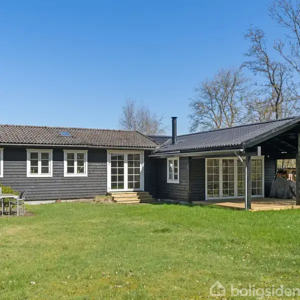 Sort sommerhus står stille i en grøn have med en overdækket terrasse til højre. I baggrunden ses træer under en blå himmel.
