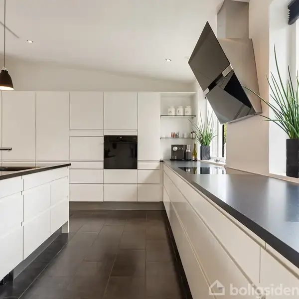 Et moderne køkken med hvide skabe og sorte bordplader. En sort indbygget ovn er synlig. Store vinduer slipper lys ind. Grønne planter pynter i vindueskarmen.