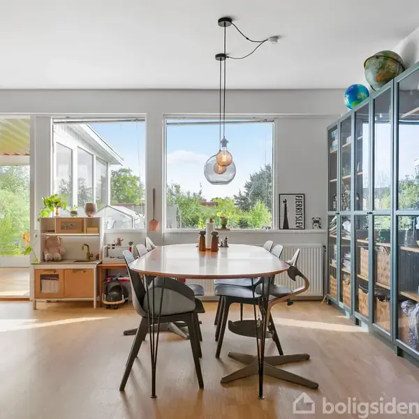 Spisebord med fire stole står i et lyst rum med trægulv. Store vinduer bagved viser en have. En glasmontre til højre indeholder bøger. En lampe hænger over bordet.