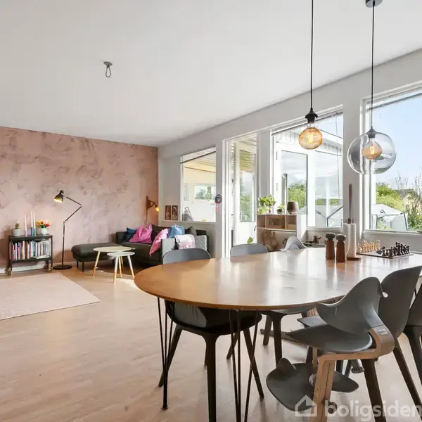 Spisebord med sorte stole står i en lys stue. Ved siden af er en sofa med puder ved en pink væg. Store vinduer giver udsigt til en terrasse med planter.