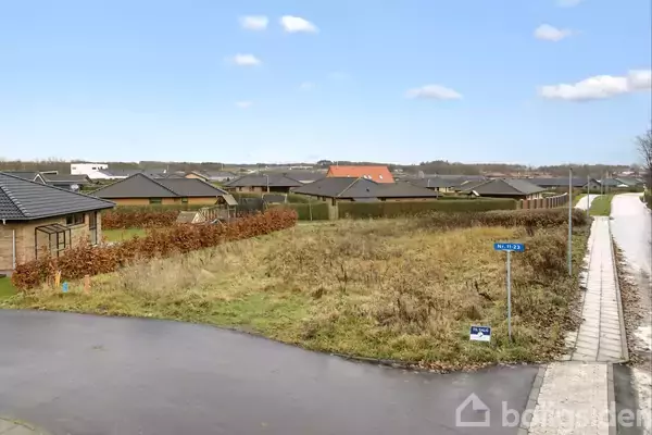 Tomt med buske og græs ligger mellem villahuse. Asfalteret vej og fortov danner en ramme om området. Skilt med teksten "Solsikkevej" står på græsset tæt ved vejen.