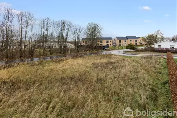 En åben græsmark på en stille vejkant med moderne boliger i baggrunden og træer langs vejen under en klar blå himmel.