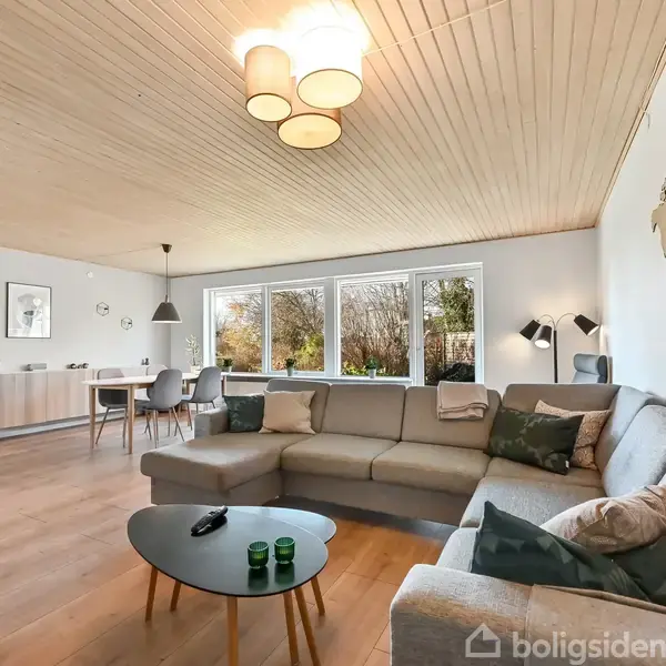 Stor hjørnesofa står i en lys stue med træloft og trægulv, omgivet af store vinduer, der giver udsigt til en have. Kortmotiv på væggen.