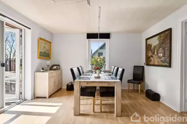 Et spisebord med seks stole står i et lyst rum. Bordet er dækket med tallerkener og blomster i midten. Vægge har kunstmalerier, og der er store vinduer med udsigt.