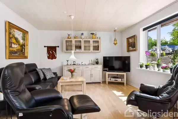 Sort lædermøblement omgiver et sofabord i en lys stue. Malerier og dekorative genstande pryder væggene, og et stort vindue lader sollys strømme ind, med udsigt til en have.