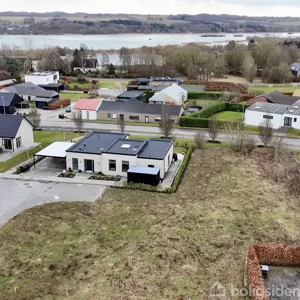 Et moderne hus med fladt tag står fredeligt i et beboelseskvarter omgivet af andre huse og grønne områder, med en fjord i baggrunden under en overskyet himmel.