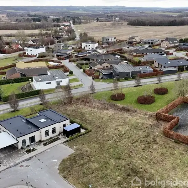 Bebyggelse med parcelhuse ligger langs en vej på landet. Huse varierer i størrelse og stil, omgivet af haver og græsområder. Marker og åbne vidder strækker sig i baggrunden.