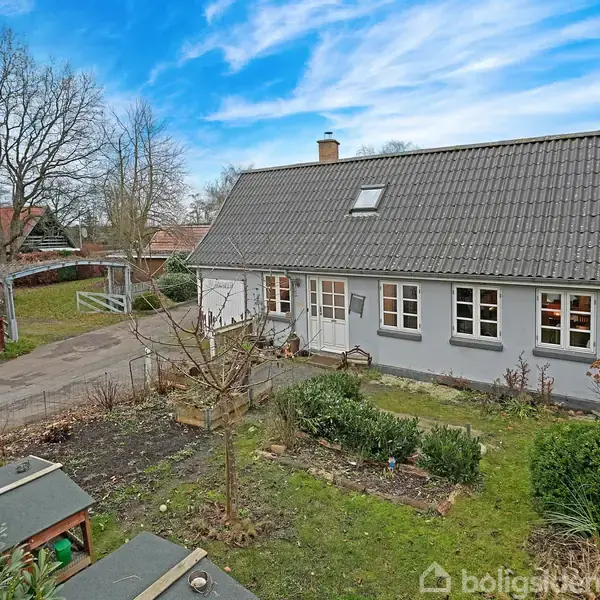 Et hvidt hus med skråtag står i en have med buske og stier, mens en blå himmel med lette skyer danner baggrund. En rød bygning ses til højre.