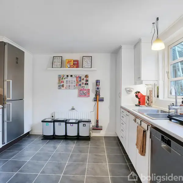 Køkken med hvide skabe, grå fliser og et sort køleskab. En kaffemaskine står på bordet, tre affaldsspande ved væggen, og vinduer giver naturligt lys. To plakater hænger på væggen.