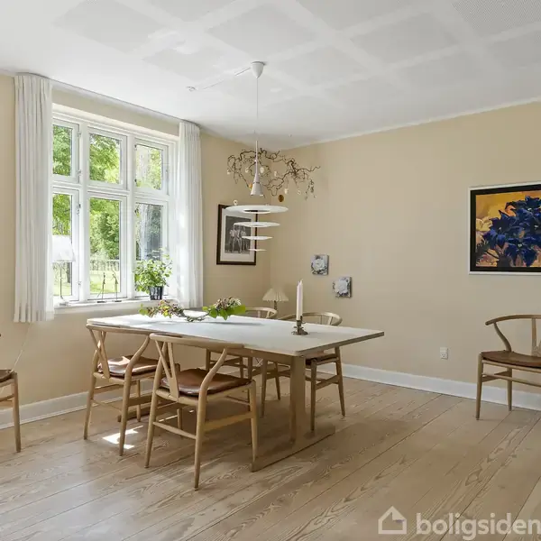 Et lyst spisebord med stole står i et rum med trægulv. Bordet er pyntet med blomster og dekorationer. Væggene har moderne kunst, og et stort vindue slipper lys ind.