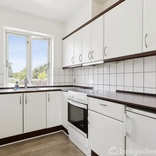 Hvid facadebygning med rødt tegltag, vinduer og hoveddør ses ved fortov. En bil parkeret foran bygningen. Skygger fra lyktestolpe falder på væggen.