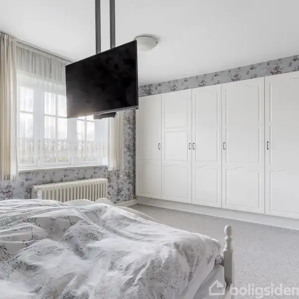 Et soveværelse med en stor seng dækket af mønstret sengetøj, en vægmonteret fladskærms-TV, og hvide skabe i baggrunden. Blomstret tapet og gardiner på vinduet.