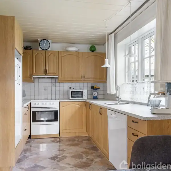 Køkken med træskabe og hvidevarer, herunder ovn og opvaskemaskine, placeret i et lyst rum med flisebelagt væg og stor vinduesudskæring. Spisebordsstol ses i forgrunden.