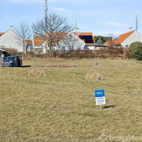 Græsmark med et skilt fra "Realmæglerne" tæt på en bil. Baggrunden viser flere huse med røde tegltage, omgivet af enkelte træer og buske.