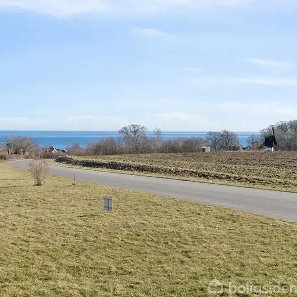 Græsplæne strækker sig langs en asfalteret vej; havet ses i horisonten med få træer og bygninger. Himmel dækker landskabet.