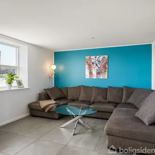 En stor, grå sofa står i hjørnet af en stue med turkis væg. Et rundt glasbord står foran sofaen. Rummet har flisegulv, vægmaleri og et TV på væggen.