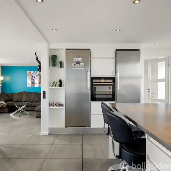 Køkken med spisebar og to sorte stole, i et moderne hjem med åben adgang til stue. Stuen har en blå væg, brun sofa og moderne indretning.
