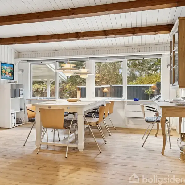 Et spisebord med otte stole står i et lyst rum med træbjælker i loftet. Store vinduer giver udsigt til en have. Møblerne inkluderer også en skænk og lamper.