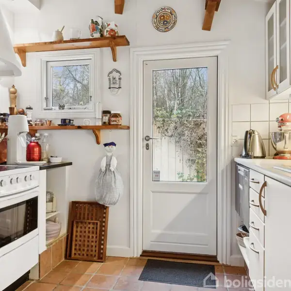 Et lyst køkken med en elektrisk komfur står forrest og en dør med glasåbning viser træer udenfor. Hylder med krydderier og køkkengrej omkranser rummet.