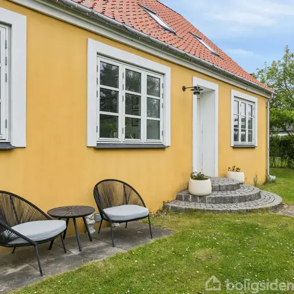 Gul husfacade med to sorte stole og et lille bord foran. Trapper fører til en hvid hoveddør. Haven har græsplæne og træer, og himlen er skyfri.
