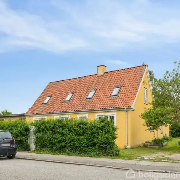 Et gult hus med rødt tegltag har flere tagvinduer. Det står i en grøn have med træer og buske. En sort bil er parkeret på vejen foran huset.