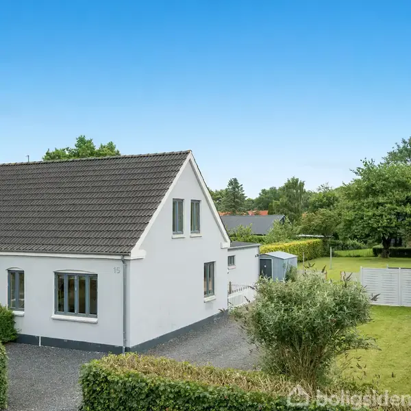 Et hvidt hus med skråtag står stille. Omkring det er en velplejet have med hække og en grusvej. Baggrunden har flere træer under en blå himmel.