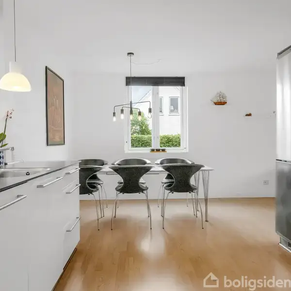 Grønt hjørnesofa står i stue med trælaminatgulv. Ved siden af er et hvidt sofabord med en plante. Væggen har billeder, og store vinduer giver udsigt til haven.
