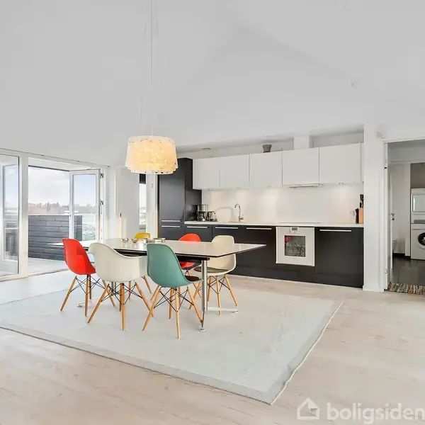 Denne moderne spisebord med farverige stole står på et tæppe, overbelyst af en loftslampe. Bagved ses et åbent køkken med hvide og sorte elementer, i en lys stue med store vinduer.