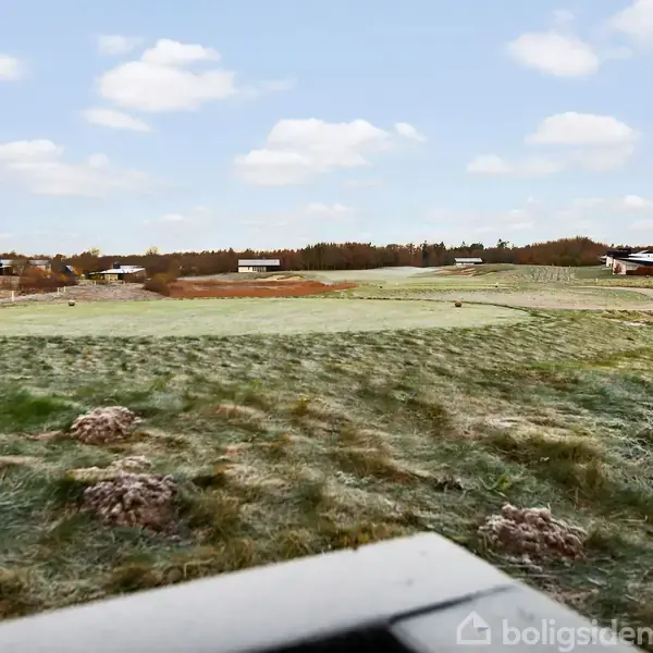 En åben mark med spredte buske under en blå himmel med skyer. I baggrunden ses enkelte bygninger langs horisonten. Markens græs ser frostbelagt ud.
