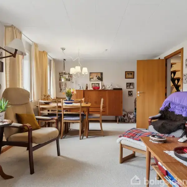 En stue med beige tæppe og lyse gardiner. Møbler inkluderer borde, stole og lænestole. Væggene er dekoreret med billeder og hylder. Rummet er lyst og hyggeligt.