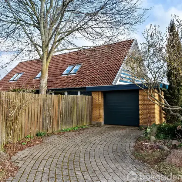 Et rødt murstenshus med et skråt tag har en lukket garageport. Huset er omgivet af et hegn, beliggende i en have med træer og buske.