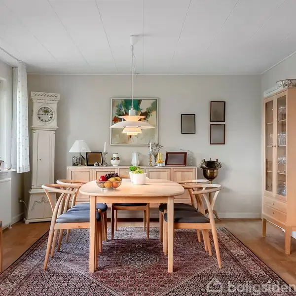 Spisebord med seks træstole står på et orientalsk tæppe. Frugtskål og lampe ovenover. Omgivet af en skænk, vitrineskab og stolur i en lys stue.